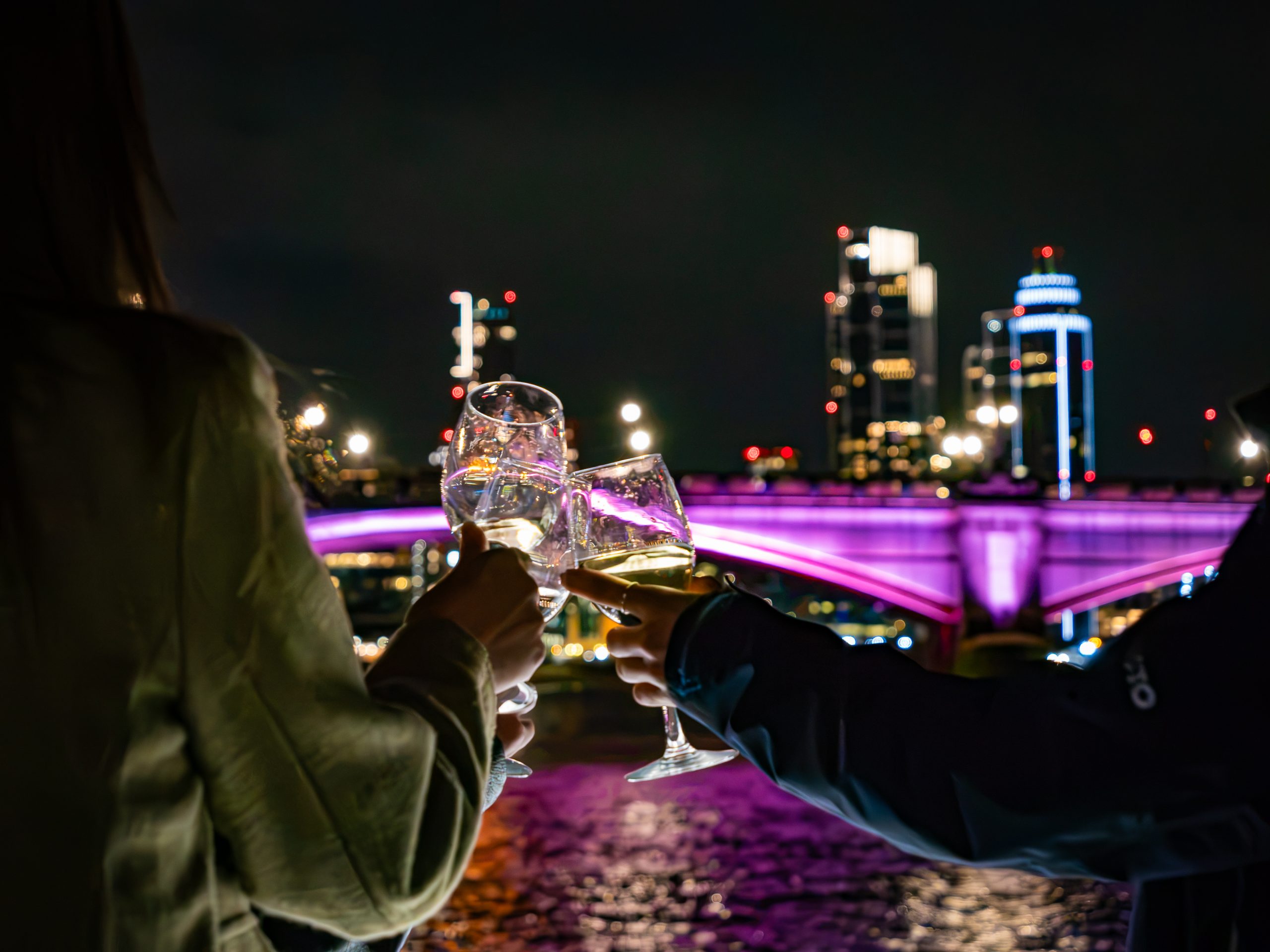 Three people toasting with wine glasses in front of a lit bridge at night.
