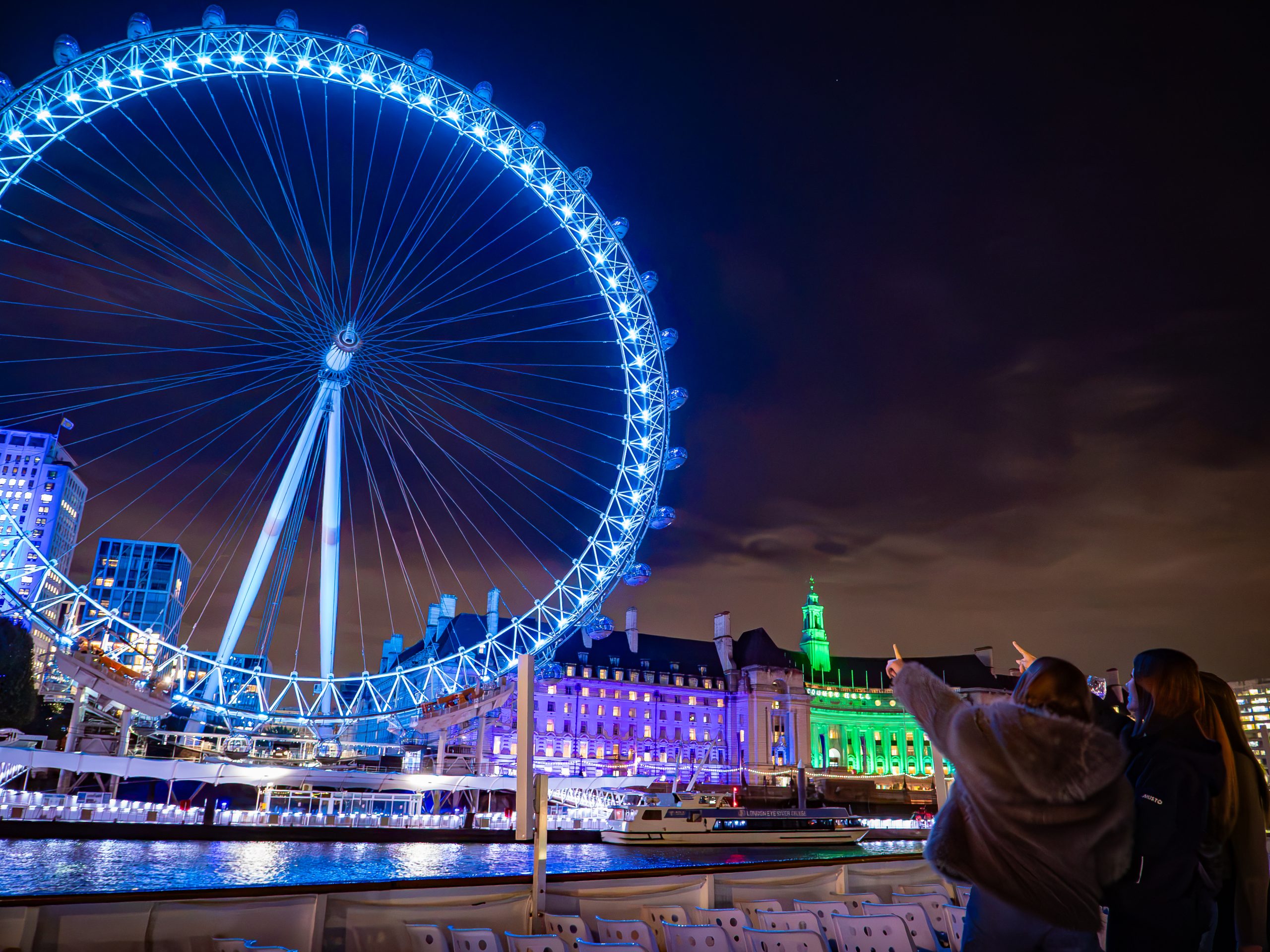 Illuminated Ferris wheel at night with people pointing at it in the foreground.