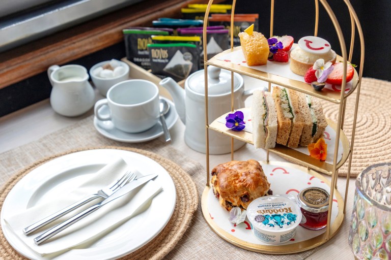Three-tier tray with sandwiches, scones, and desserts for tea, with cups and condiments on a side table.