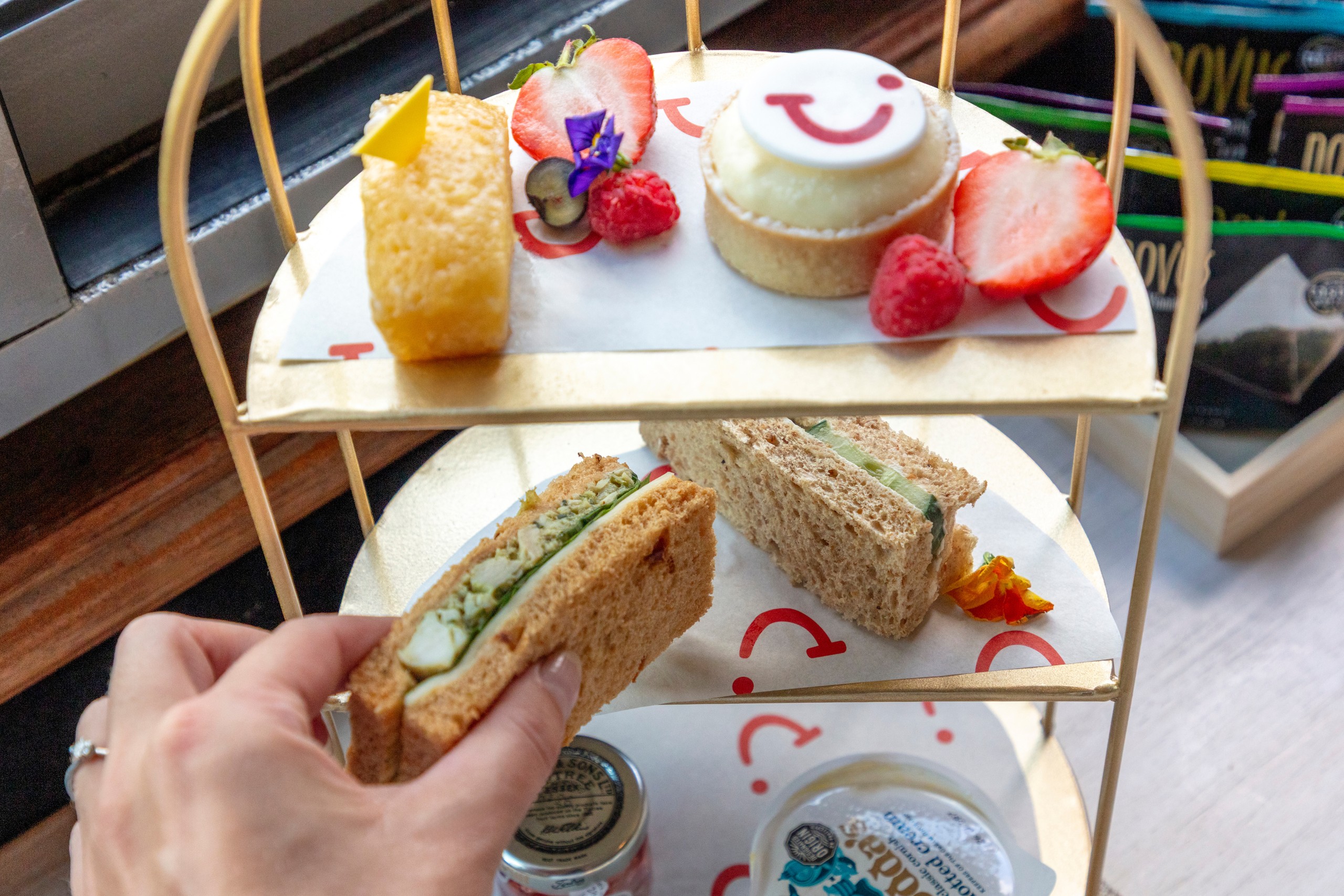 Tiered tray with desserts and sandwiches, hand reaching for a cucumber sandwich.
