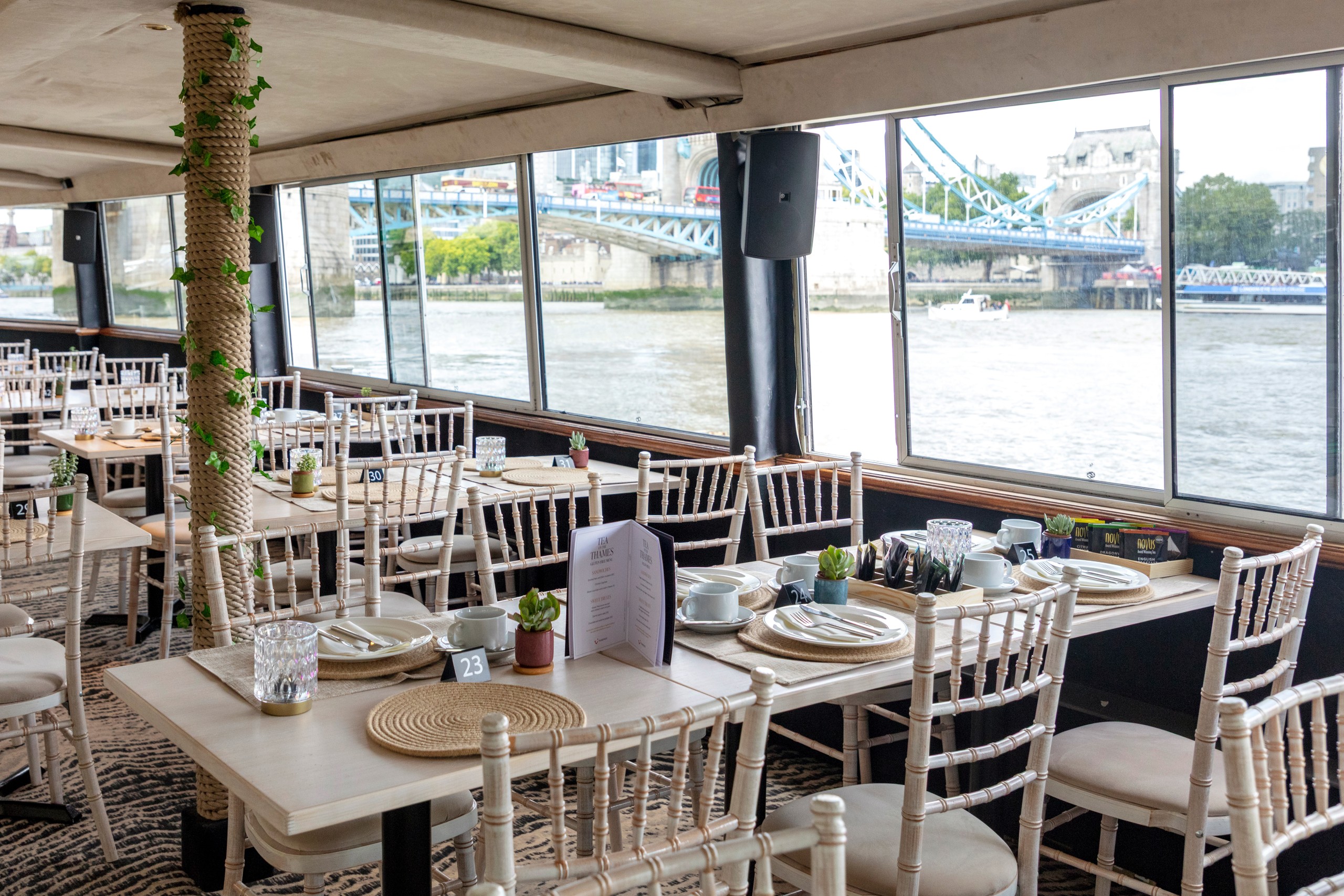 Inside a riverside restaurant with tables, chairs, and large windows showing a bridge view.