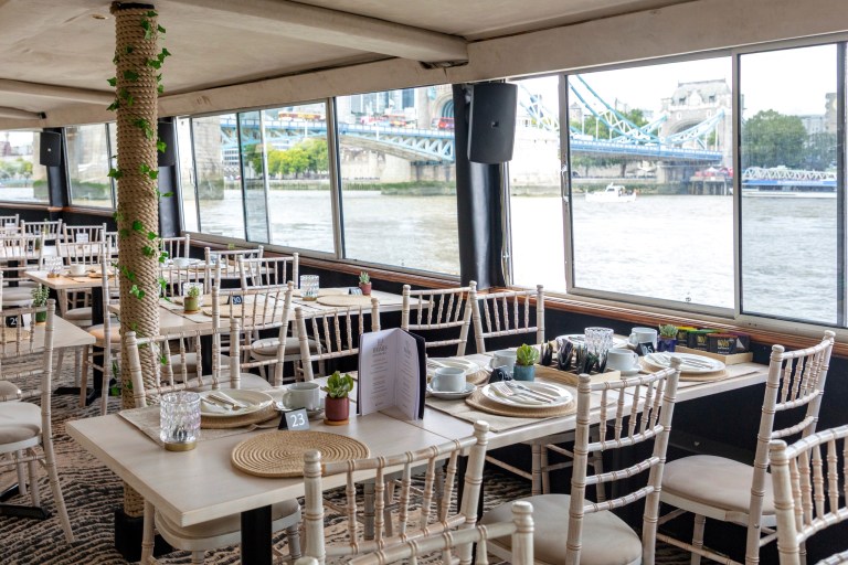 Inside a riverside restaurant with tables, chairs, and large windows showing a bridge view.