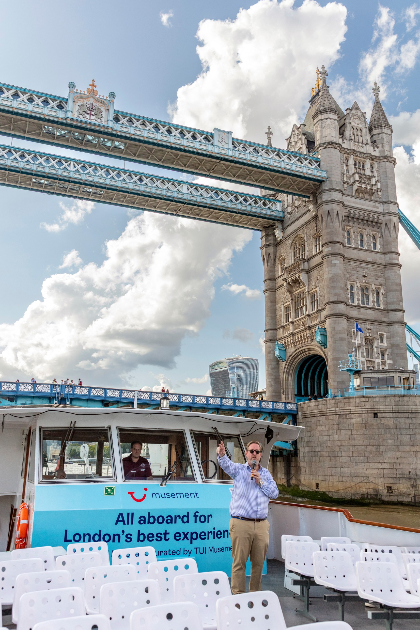 Man speaking on a boat near Tower Bridge, London, with cloudy sky.