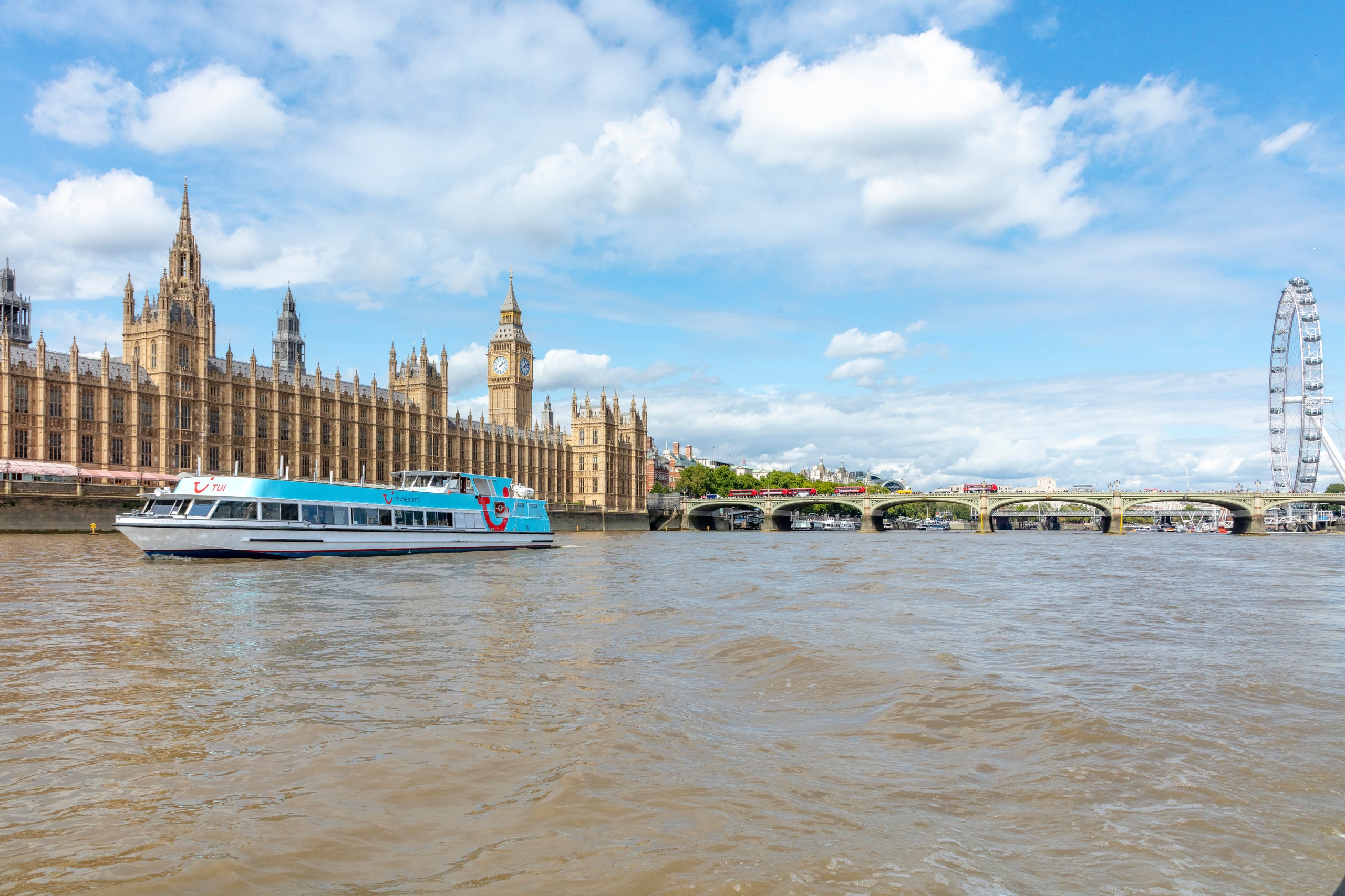 Riverboat on Thames with Big Ben and London Eye in background under blue sky.
