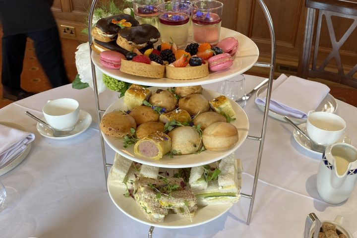 Three-tier afternoon tea stand with cakes, sandwiches, and desserts on a table with tea cups.