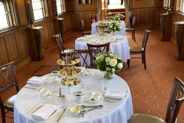 Elegant dining room with round tables set for afternoon tea, white tablecloths, and wooden paneling.