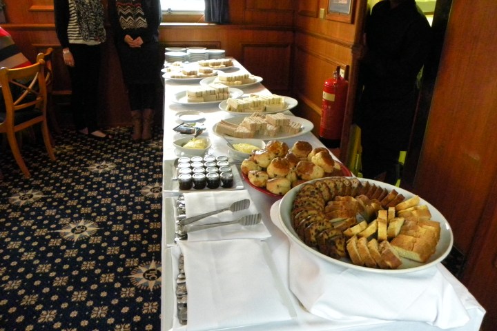 A buffet table with various breads, pastries, scones, and condiments. Three people stand nearby.