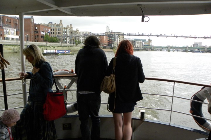 People on a boat deck looking at river scenery with city buildings in the background.