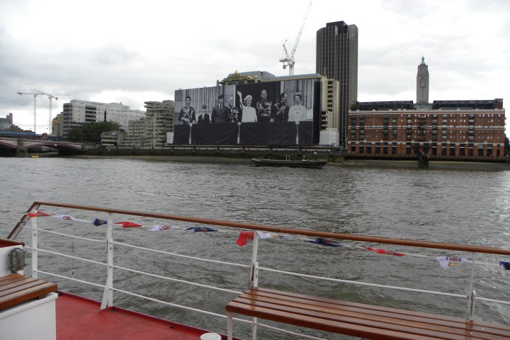 River view with large mural on building, benches on boat deck, and cranes in the background.