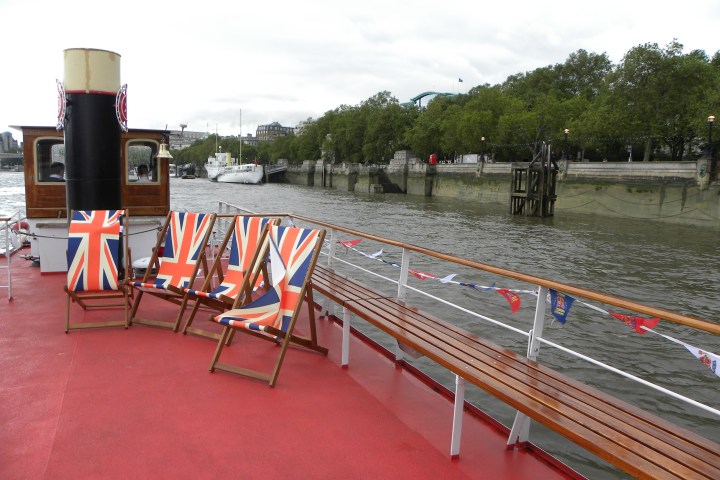 Red deck with Union Jack chairs on a boat by a riverbank and trees.