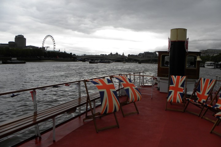 Boat deck with Union Jack chairs, London Eye and Thames River in the background under cloudy sky.