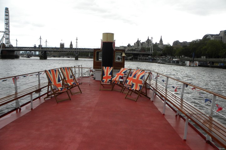Back deck of a boat with Union Jack chairs, river view, and skyline including a Ferris wheel and bridge.