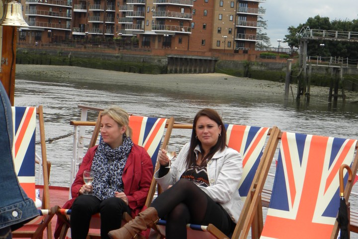 Two people sitting on Union Jack deck chairs on a boat near waterside buildings.