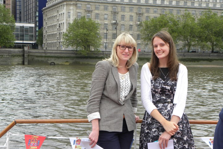 Two women standing by a riverside with a large building and trees in the background.