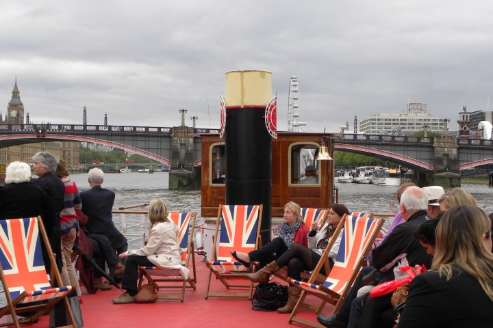 People seated on a boat with Union Jack deckchairs on a river near a bridge and Big Ben.
