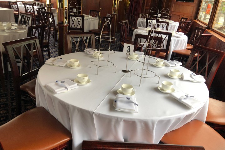 Dining area with round tables, white tablecloths, teacups, and tiered stands, prepared for guests.