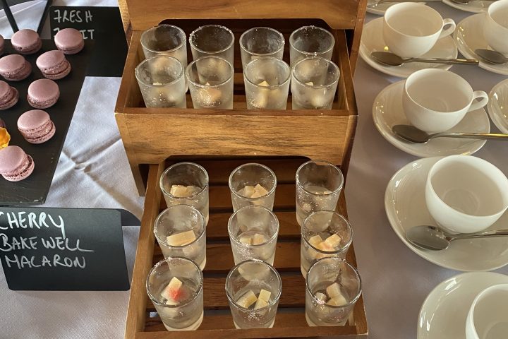 Small glasses of wine jelly on wooden tiers beside white teacups and purple macarons on a table.