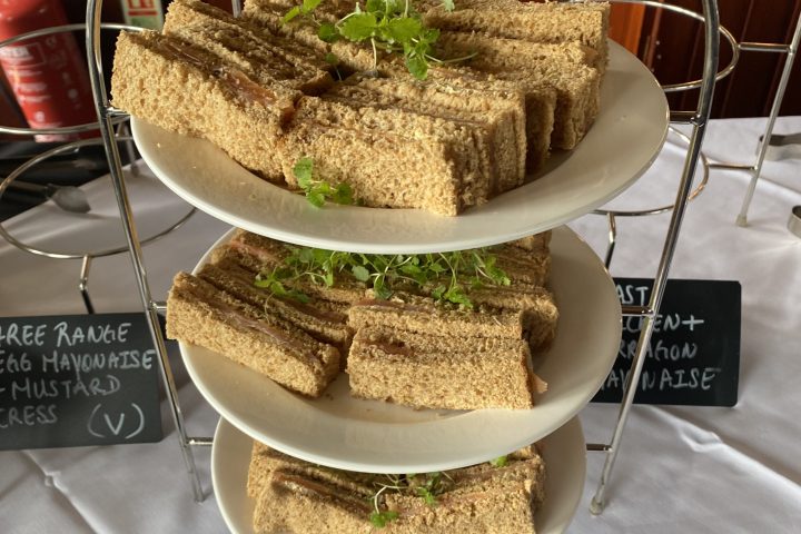 Three-tier stand with assorted sandwiches on white plates, garnished with herbs, displayed on a white tablecloth.