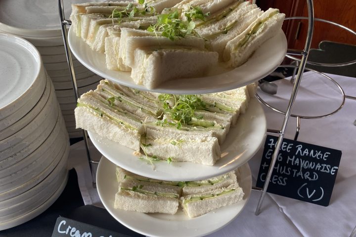 Tiered stand with cucumber sandwiches and handwritten signs on a table with stacked plates nearby.