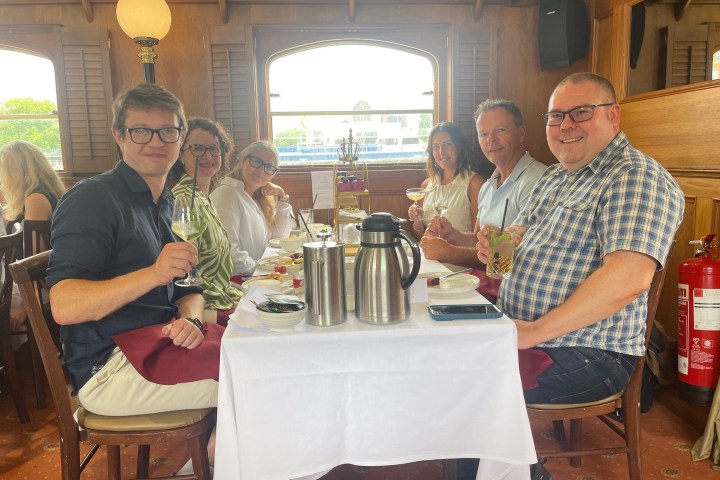 Six people sitting at a dining table with drinks on a boat.