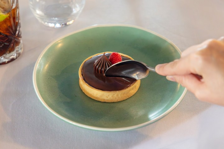 Hand using spoon on chocolate tart with raspberry on green plate.