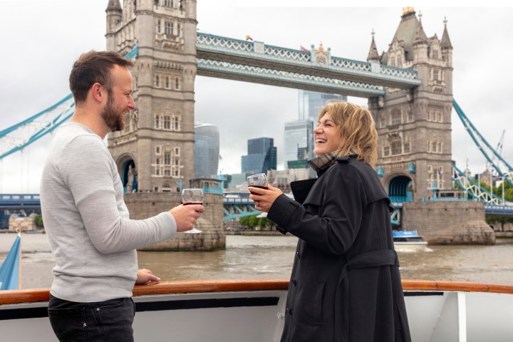 Two people on a boat holding drinks, with Tower Bridge in the background.